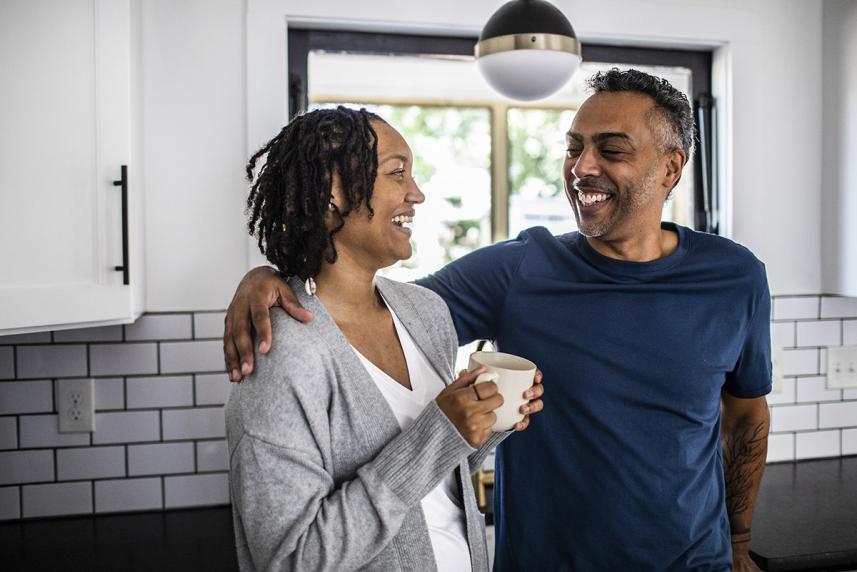 A man with his arm around his partner's shoulder while they smile and enjoy a cup of coffee