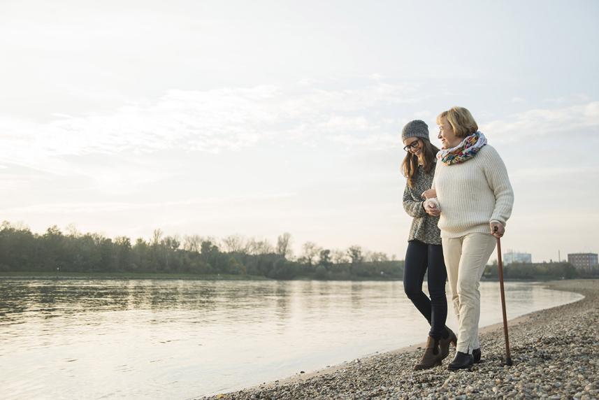 An adult dauther walking on a beach with her senior mom and holding onto her arm. 