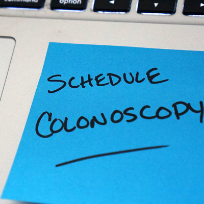 A posit note stuck to a laptop. It reads, Scheudle Colonoscopy
